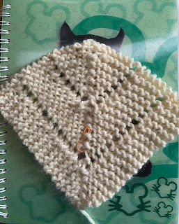 A bit of cream colored knitting on a circular knitting needle. The knitting is mostly garter stitch with some eyelets forming Vs in the triangular piece. The top edge of the triangle bows in the center rather than laying straight.