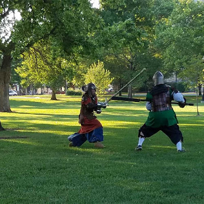 Two people in medieval armour in a grassy, sunny park with large trees. The taller person, in a green tunic, is levelling a polearm at the shorter, red-coated person, who is swinging a greatsword up and taking a step sidways.