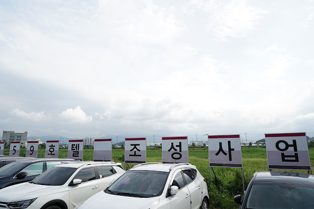 넓은 초지 앞에 차량 몇 대가 주차되어 있고 차량과 초지 사이에 '59 호텔 조성 사업'이라고 적힌 대형 표지판이 세워져있다.