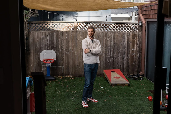 A man in a white sweater stands in a yard before a wooden fence