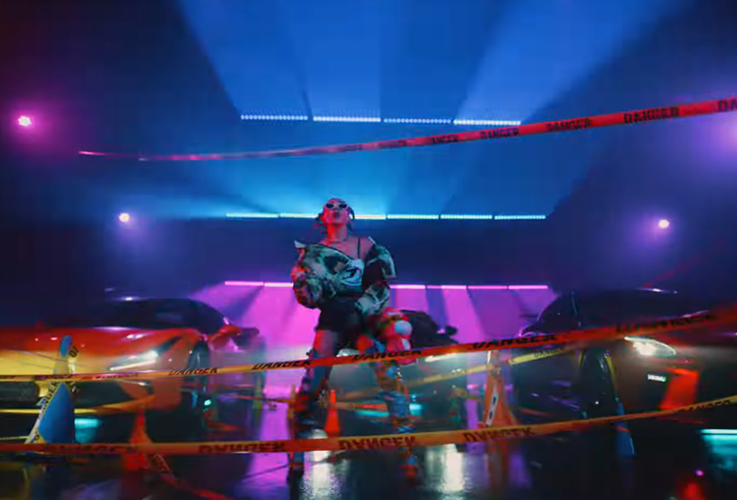 A screenshot from Koda Kumi’s music video for “Vroom”. Featuring her in a warehouse with blue and purple lights, sports cars and danger tape strewn everywhere.