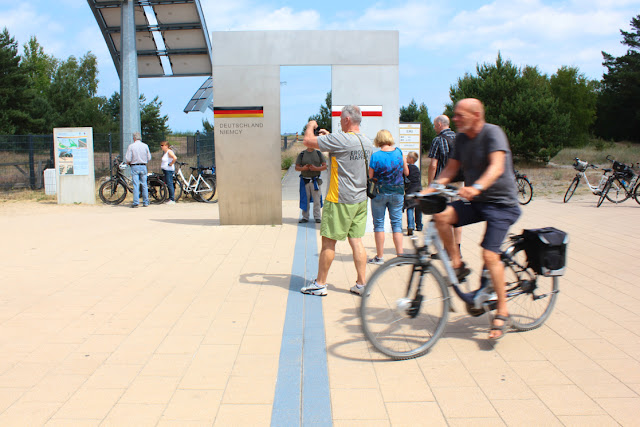 Na polsko-niemieckiej granicy między Świnoujście a Ahlbeck. Ścieżka rowerowa z namalowaną na niej niebieską linią, znaczącą granicę państw. Nad linią stoi znak, znaczący polską i niemiecą stronę, Przy znaku stoi wiele rowerów. Kilka osób fotografuje przejście. Na rowerze przejeżdża graniczną linię starszy mężczyzna.