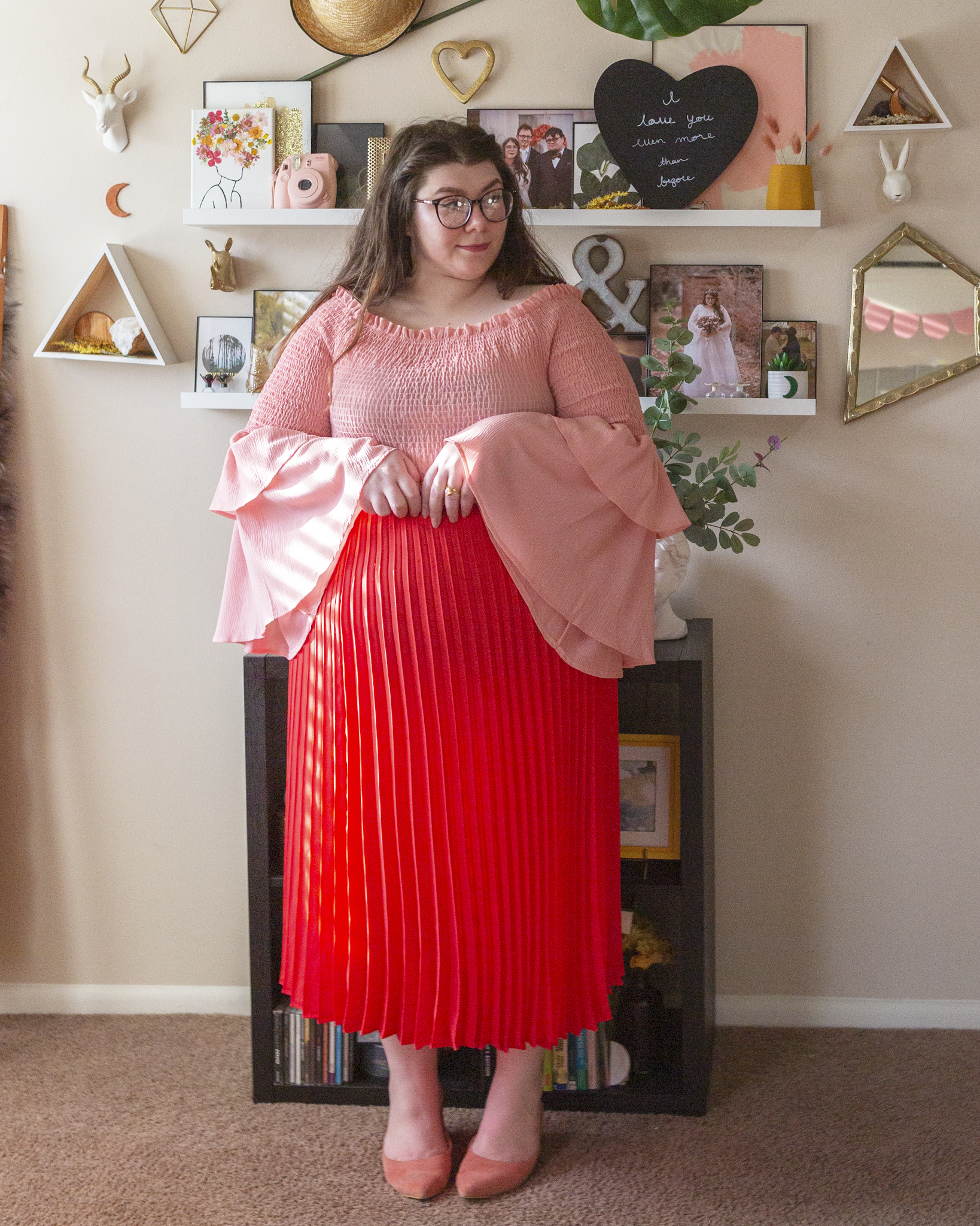 An outfit consisting of a pastel pink shirred three tiered off the shoulder blouse tucked into a coral pink pleated midi skirt and pastel pink heeled clogs.