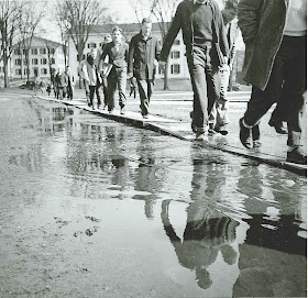 A series of men walking along boards to avoid standing water on the ground.