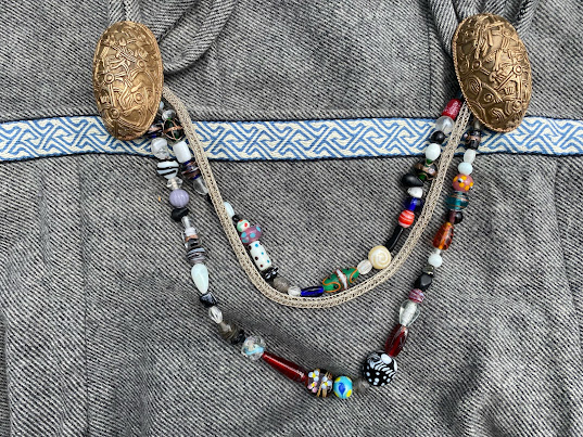 A close-up of a grey wool Viking apron dress with blue and white tablet woven trim and two oval shaped brass brooches. Two strands of beads and a silver chain are strung between the brooches.