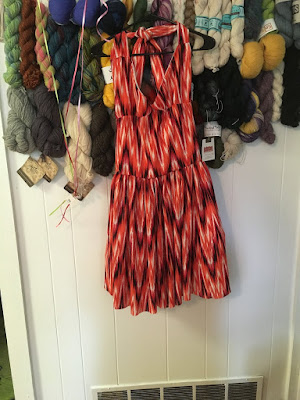 A halter-top sundress in coral, black, and white fake-ikat fabric, hanging in front of a white wall with dozens of skeins of yarn clustered in rows near the top.