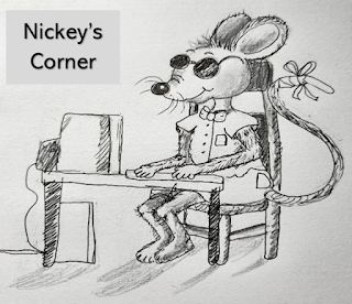 Nickey, one of the Blind Mice, is seated at a table in Nickey's Corner with his front paws on a computer keyboard. He is wearing a short sleeve shirt and shorts with a bowtie and sunglasses. The tip of his tail is bandaged.