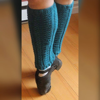 A pair of teal legwarmers with cable and textured panels around them on a dancer wearing Irish hardshoes and standing en pointe, from the knees down.