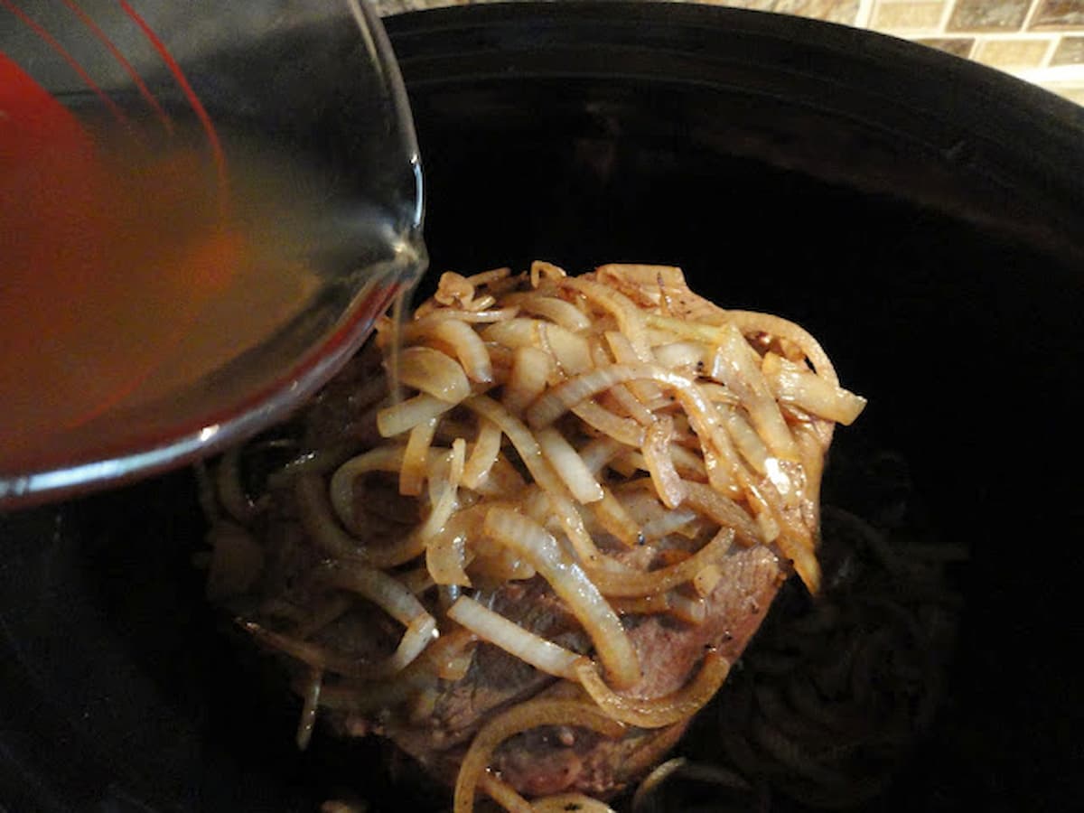 Beef Broth being poured over caramelized onions and browned beef chuck roast in a slow cooker.