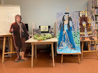 Dzień Czytania Tolkiena_2 Na tle ściany stoją w szeregu: portret Hobbita umieszczony na sztaludze, postać Biblo Bagginsa, stolik w książkową wystawką, 2 postaci typu fotobudka: elfica i krasnolud.