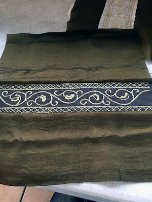 A band of gold floral embroidery on brown fabric, dividing a piece of green-gold fabric horizontally in half.