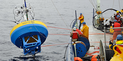 On the right side of the image is a group of at least 6 people, wearing safety vests and helmets, on the deck of a research vessel holding ropes attached to a surface mooring on the left of the image. The surface mooring is approximately 6-7 times the height of the people, is slightly tilted away from the camera, and has a spindly white top over a blue painted base. Around the middle of the mooring is a yellow band with the WHOI and OOI logos visible in blue letters.