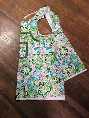 A photo from above of an apron made of green, turquoise, and grey tropical floral patterned fabric, folded vertically in half at the center front, with the straps slightly crumpled from crossing each other. A large, rectangular pocket with a quarter-circle cut from the top corner is placed about five inches below the top of the bib, with a smaller square pocket centered on it. The smaller pocket is completely edged in light lime-green fabric with densely scattered gold dots, and the larger pocket is only edged at the top. The main sections of the apron have white selvedges at the hems, and are mismatched by about five inches, forming a ragged down-pointing arrow.