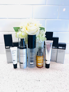 Five PCA Skin products arranged neatly on a marble countertop, including C & E Advanced, ExLinea Peptide Smoothing Serum, ProMax Age Renewal, Hyaluronic Acid Boosting Serum, and Vitamin B3 Eye Brightening Cream. The bottles are a mix of vibrant colors with sleek designs, reflecting the brand's elegant and clinical aesthetic.