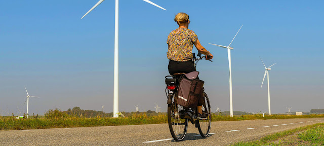 Una mujer pasa en bicicleta junto a unos turbinas eólicas en una carretera rural de Heijningen (Países Bajos).   Unsplash/Les Corpographes