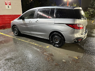 A silver mazda 5 parked outside a dealership in the rain.