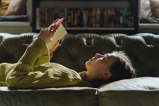 photo of a young woman laying down on a couch reading a book.