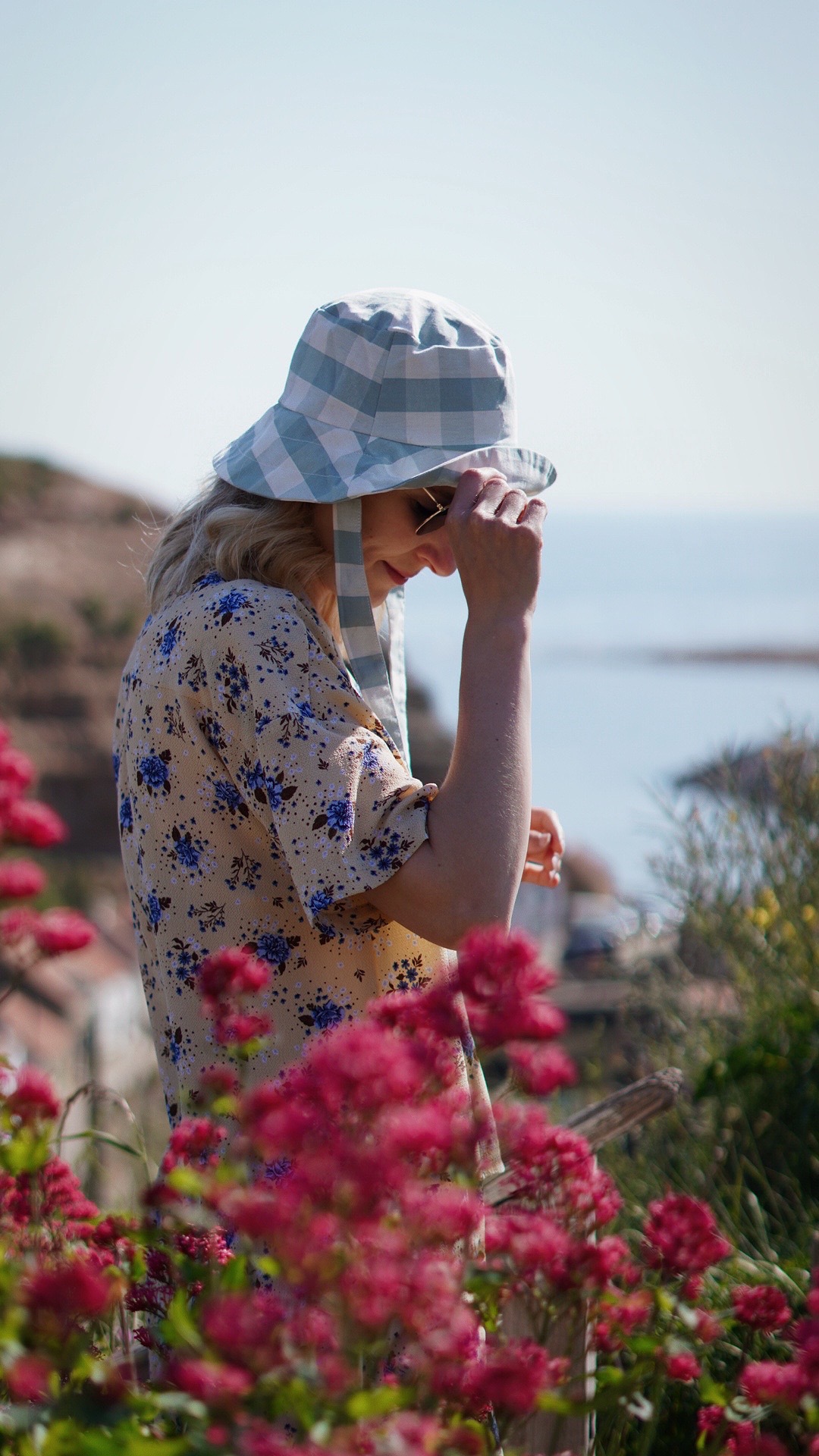 Amy is standing in front of a view of Staithes, North Yorkshire. She has her hand up to her blue gingham sun hat and is wearing a yellow blouse. There are flowers in the foreground