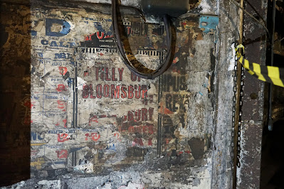 Photograph of dirt-stained, tattered theatre posters. Fragments of the names of Shakespeare plays are visible, and the words 'Tilly of Bloomsbury' are large and legible in the centre.