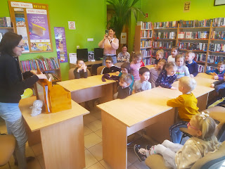 Pani bibliotekarka opowiada dzieciom bajkę. Dzieci siedzą przy stolikach. W tle Pani przedszkolanka i regały z książkami.