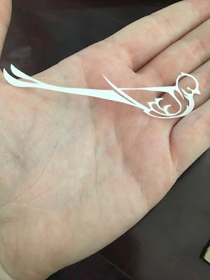 A delicate, stylized silhouette of a flycatcher cut from white paper, lying across a white hand. The cutout just reaches from the base of the thumb to the base of the pinky, across the palm.