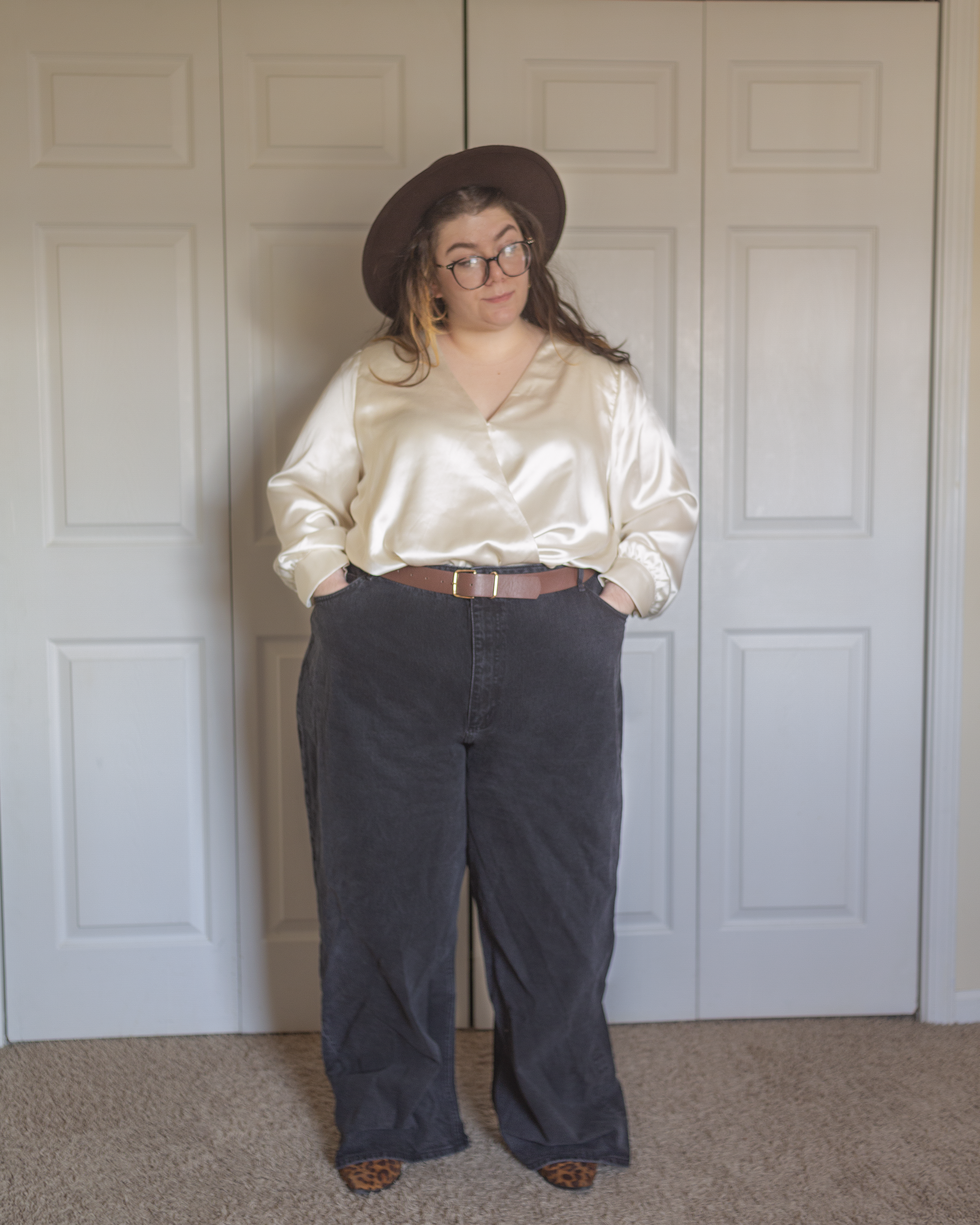 An outfit consisting of a wide brim dark brown fedora, a cream colored long sleeved satin blouse tucked into wide leg black denim jeans and brown and black leopard print boots.