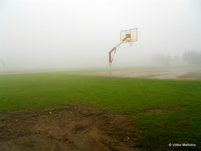 The World's Highest Cricket Ground, Chail. When seen from a purely tourist perspective, Chail doesn't have too much to do. However there are several hidden treasures that are worth exploring. One of them is the World's highest cricket ground. I have a suspicion that the place would have been quite dull if the weather wasn't so magical. The board indicating that this is indeed the world's highest cricket ground at 7500 feet from the sea level. The cricket ground is used by the Chail Military School. The ground was built in 1893 and is still frequently used and well-maintained. My family on the stairs leading to the Cricket Ground. Clouds had started gathering by now and the ground was barely visible. The cloud-curtain lifted for a second to allow us a view of the ground. We were surprised to see a basketball court as well. It is surprising then that this isn't the world's highest basketball court as well. A quick search on the Internet didn't reveal which basketball court gets the honour. And it is also worth mentioning here that this  ground is also used to play polo during the vacations. A beautiful bald tree with a tree-house. You need to walk around the cricket stadium a bit to reach this point. The cricket ground was on higher grounds when compared to the road we were walking on. I wish I had been able to click a better angle though. The inactive scoreboard. Honestly, when I was told about this cricket ground, I was expecting a stadium and not really just a ground. But anyways, the visit was worth the time so no complaints. The expanse of the playground surrounded by tall deodars. The light clouds drifting through these giants make the entire environment quite haunting. Playing cricket here would be so much fun. I wonder how many balls were hit too hard and lost to the game forever in the dense foliage surrounding the ground.