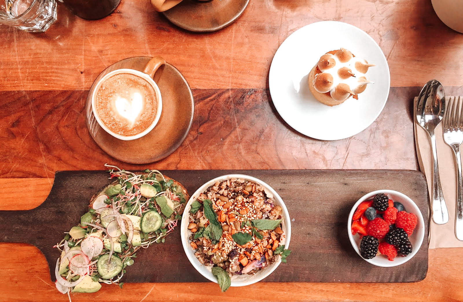 sur une table en bois du restaurant république à los angeles sur une table en bois du restaurant blu jam café à melrose avenue à los angeles un cappuccino avec un coeur de dessiné à l'intérieur et devant une planche en bois avec un avocado toast à côté un bol de muesli avec des feuilles de menthe et quelques fruits rouges dans une coupelle et en haut à gauche une petite tarte au citron meringuée