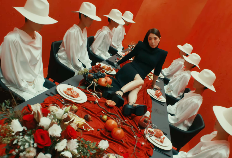 BoA lying on a dining table, wearing a black dress, as women in white dresses and white wide brimmed hats sit around her.