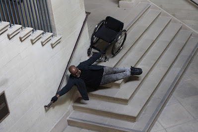 A brown-skinned large-bodied black person with shaved head & beard, wearing glasses, black shirt and grey jeans, with one hand on a rail and the other holding a wheelchair, seemingly floating above a set of stairs.
