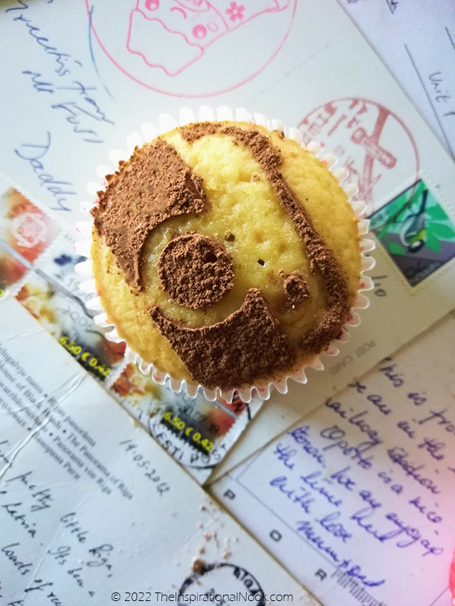 Camera stenciled with cocoa powder on a yellow cupcake with a background of postcards