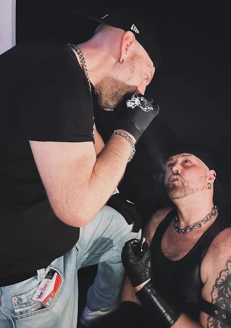 Two scally lads blowing smoke on each other wearing backwards baseball hats, black t-shirts, blue jeans and black leather gloves