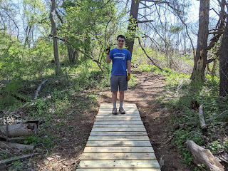 Teenage boy standing on a newly constructed, short wooden bridge in the woods. The boy is holding a hammer and wearing work gloves.