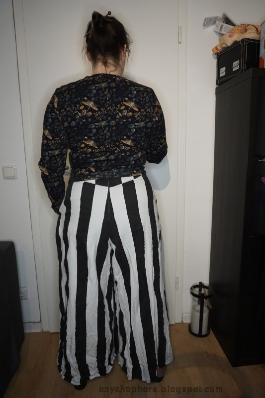 Rückansicht einer stehenden Frau. Sie trägt eine Hose mit schwarz-weißen Längsstreifen und einen Pullover mit Libelle- und Blumenmotiven.