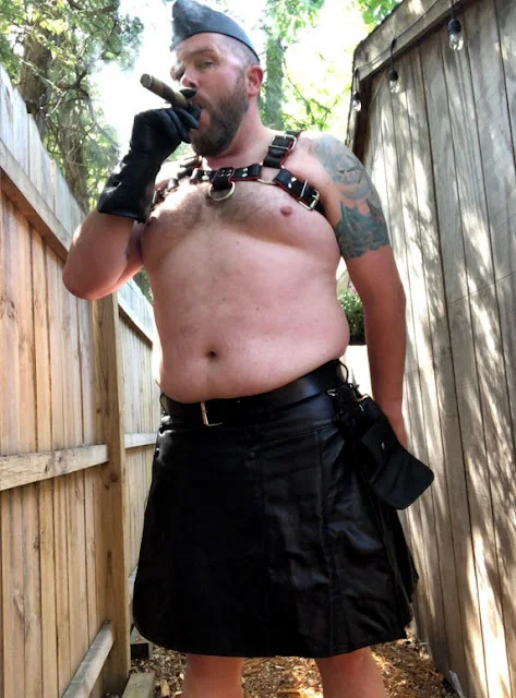 1/2 Smoking a cigar standing outside wearing leather harness,gloves, flight cap and kilt