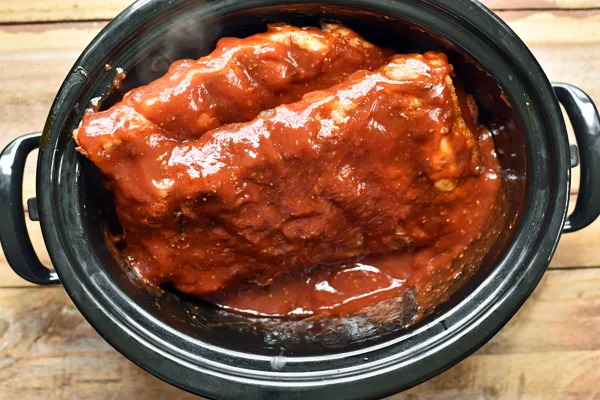 Seared St. Louis style ribs in a slow cooker, smothered in homemade BBQ sauce. The meat is seared to perfection and coated in a delicious, savory sauce, with the slow cooker resting on a rustic wooden table