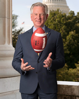 Photo of Senator Tommy Tuberville facing the camera with a football in the air between his hands and chin. Source: www.tuberville.senate.gov/about/