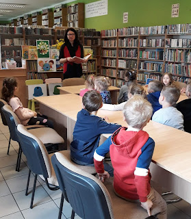 Pani bibliotekarka opowiada dzieciom o ochronie środowiska. Dzieci siedzą przy stolikach. Tło: sala biblioteczna