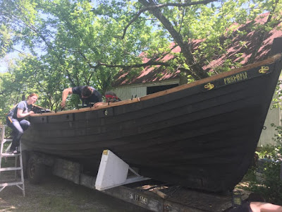 A clinker-built Viking ship on a trailer in a sun-dappled clearing, with a bright new rail clamped on, a man leaning over the rail from the inside of the boat, and a redheaded woman leaning over the rail from a ladder on the outside of the boat.