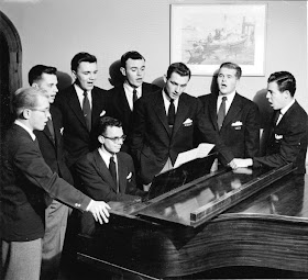A black and white photograph of men singing around a piano.