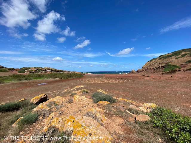A red sandstone landscape surrounds a red sandy beach in a bay surrounded by sandstone cliffs and green hills, under a blue sky with a few white fluffy clouds.