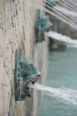 Photograph showing detail of a textured concrete wall with bronze lion heads attached, each spouting water from its mouth. One lion head fills the foreground; another is visible but blurred behind it, and more thin streams of water stream above them.