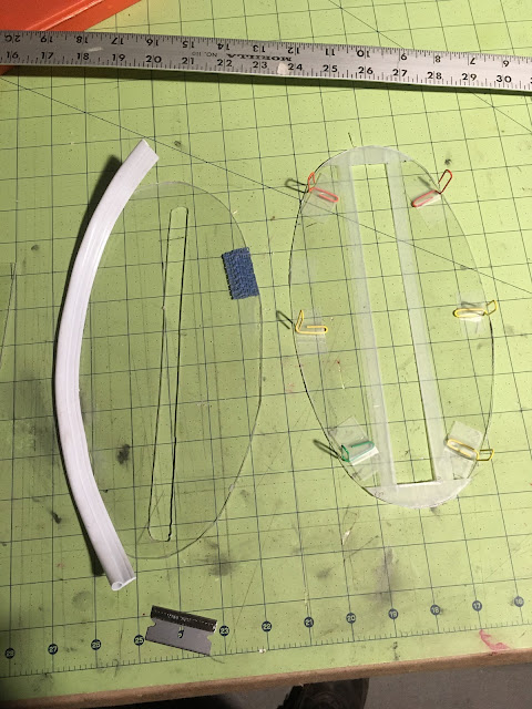 Two plexiglass oval shapes with slots on worktable. One has vinyl weather stripping glued onto edge, other has bent paper clips taped around the edge.