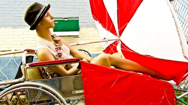 A mannequin sitting next to a patio table umbrella in a wagon that looks like a wheelchair.