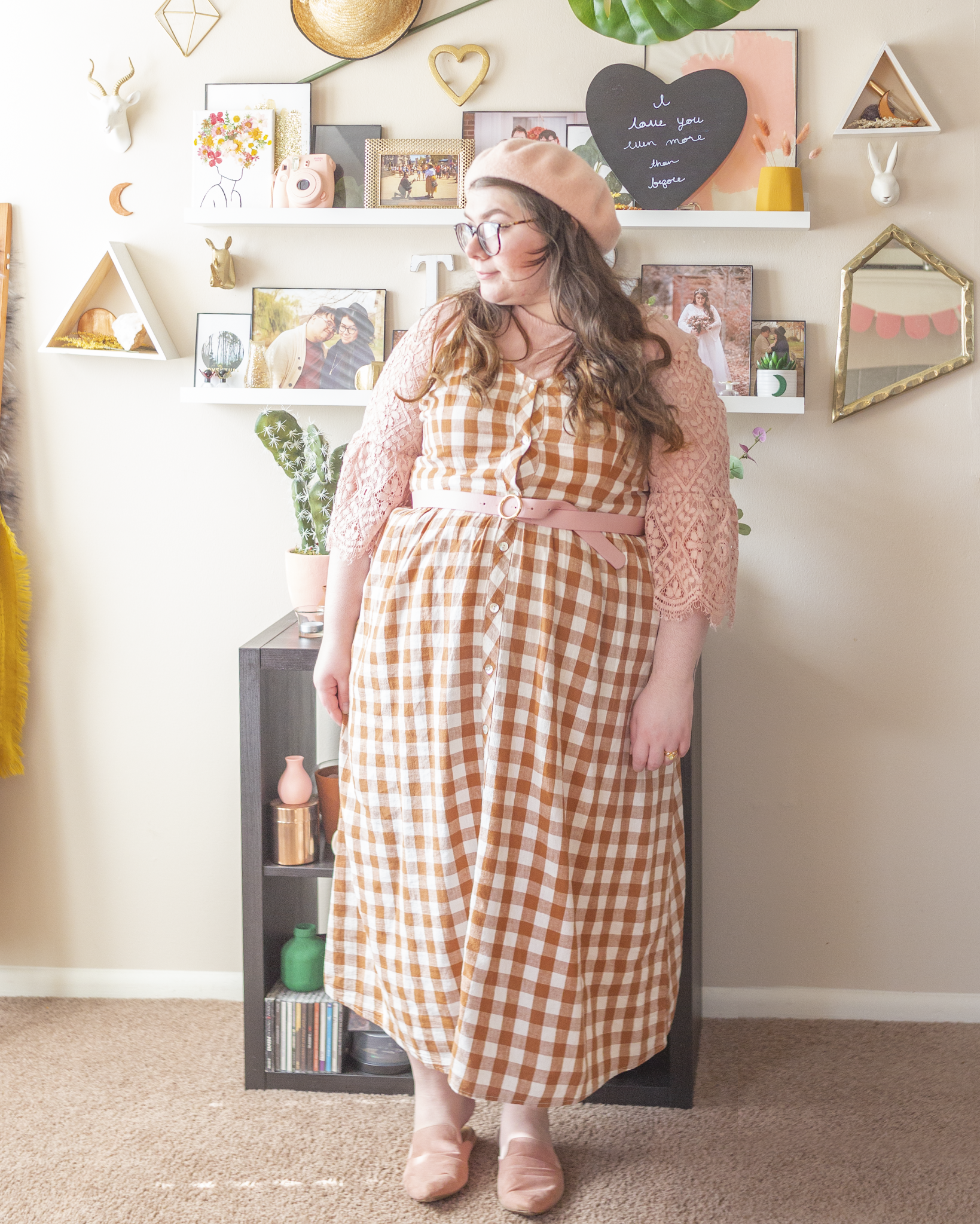 An outfit consisting of a pastel pink beret, a pastel pink midi dress with lace 3/4 sleeves under a brown gingham button down dress with spaghetti straps, belted with a pastel pink belt around the waist and pastel pink pointed toe mules.