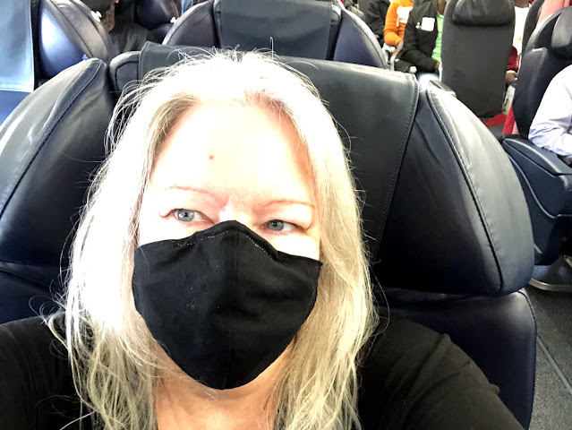 A woman wearing a black mask. She has long light hair, and is sitting in a business class seat.