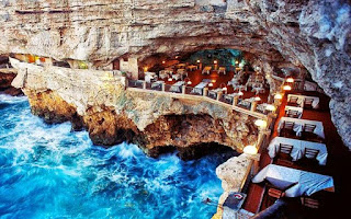 Este lugar existe e se chama Grotta Palazzese em Polignano a Mare, uma cidade situada na costa adriática da Puglia, no sul da Itália. Foto: vista aérea de um restaurante construído dentro de uma gruta: entre o paredão com saliências irregulares na parte superior, uma tímida fenda à esquerda usada como corredor e as rochas corroídas pela erosão na parte inferior, onde percebe-se as espumas brancas das ondas do mar azul anil em repuxo, surge o restaurante, à direita, em um nível acima cinco mesas retangulares, toalhas brancas, em cada coluna baixa quadrangular da mureta, há uma luminária com cúpula arredondada que remete a um cogumelo, iluminação delicada sem interferência na arquitetura natural; ao fundo, em um nível mais baixo, um amplo vão protegido pela rocha em arco, mesas, cadeiras e luminárias no mesmo estilo compõem o espaço e criam uma atmosfera romântica.