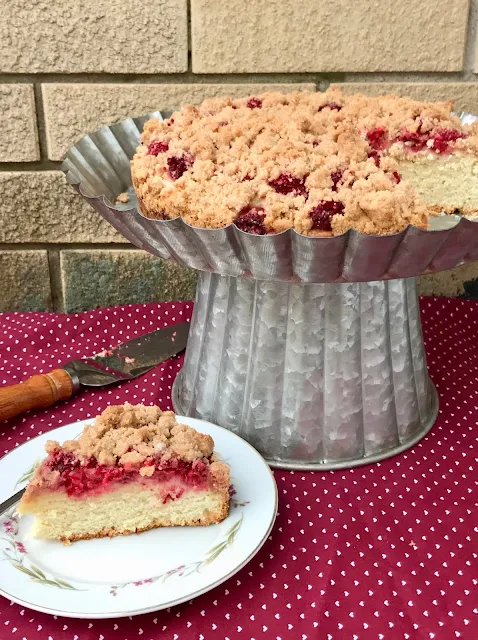 Side view of a finished lemon raspberry buckle on a cake stand with a slice of cake in front on a plate.