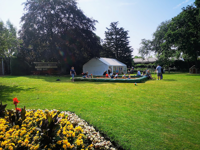 The Lawns at Bassetlaw Museum. Image shows a large turfed area of bright green grass, lots of well established trees in the distance and brightly coloured flower beds in the foreground.There is a white marquee and an extremely large sand pit being used as an artificial beach.