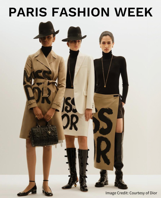 Photo of three female models wearing Dior for their Paris Fashion Week show. First model is wearing a tan trench coat with ‘MISS DIOR’ spray painted in black across the piece, black trilby hat, black pumps and a black Dior handbag. Second model is wearing a tight black turtleneck, cream blazer, cream a-line skirt with ‘MISS DIOR' spray painted in black across the piece, black knee high boots and black trilby hat. Final model is wearing a tight black turtleneck, long gold necklace, tan maxi wrap skirt with ‘MISS DIOR’ spray painted in black across the piece and black knee high chelsea boots.